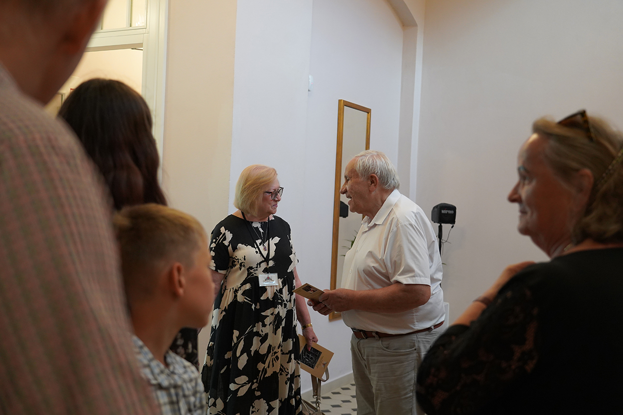 Uroczyste otwarcie wystawy, Jadwiga Inka Zamoyska z Ireneuszem Targońskim Zdjęcie przedstawia uroczyste otwarcie wystawy w oficynie północnej Muzeum Zamoyskich w Kozłówce. Grupa osób znajduje się w jasnym, przestronnym wnętrzu. W centrum kadru znajduje się starsza kobieta i starszy mężczyzna, którzy rozmawiają ze sobą z uśmiechem. Kobieta ma jasne włosy, okulary i nosi czarną sukienkę z białym, kwiatowym wzorem. Na szyi ma zawieszony identyfikator. W ręce trzyma folder. To Jadwiga Inka Zamoyska. Mężczyzna o siwych włosach i lekkim uśmiechu ubrany jest w białą koszulę z krótkim rękawem i jasne spodnie. On również trzyma w rękach ulotkę. To autor wystawy Ireneusz Targoński. Po bokach zdjęcia widoczne są inne osoby, częściowo zasłonięte – na pierwszym planie po lewej stronie chłopiec z jasnymi włosami w koszuli w kratę, obok niego osoba z długimi ciemnymi włosami. Po prawej stronie starsza kobieta w ciemnym ubraniu patrzy z uśmiechem w stronę centrum wydarzenia. Tło to jasna ściana, drzwi i lustro. W głębi widoczny jest statyw z mikrofonem lub sprzętem do nagłośnienia.