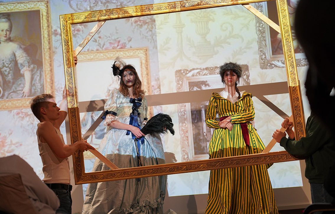 Spektakl „Tableau vivant, czyli żywe obrazy z kozłowieckiego pałacu” Na zdjęciu widać scenę przedstawienia inspirowanego dawnym malarstwem. Na pierwszym planie dwie młode osoby stoją wewnątrz dużej, złotej ramy obrazu, którą po bokach trzymają inni uczestnicy spektaklu.
Postać po lewej stronie ma na sobie błękitną, długą suknię z bufami i ciemnoniebieskimi wstążkami, na głowie czarną ozdobę z piórami. W dłoniach trzyma czarne pióro – wachlarz. Obok niej stoi osoba w żółto-zielonej sukni w pionowe pasy, z białym kołnierzem i czerwonym pasem w talii; na głowie ma futrzaną czapkę.
W tle wyświetlone są powiększone reprodukcje obrazów przedstawiających kobiety w strojach z XVIII wieku w złoconych ramach. Całość tworzy wrażenie, że aktorki stały się częścią ożywionego obrazu.
Scena jest barwna i teatralna – światło skupia się na postaciach w ramie, podkreślając efekt „żywego obrazu”.