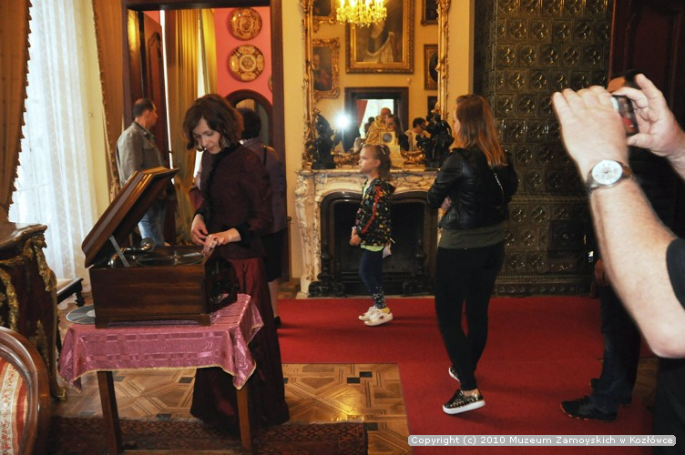 Fotografia. Kobieta ubrana w stylizowaną bordową suknię nastawia gramofon. W tle spacerujący po pomieszczeniu turyści.
